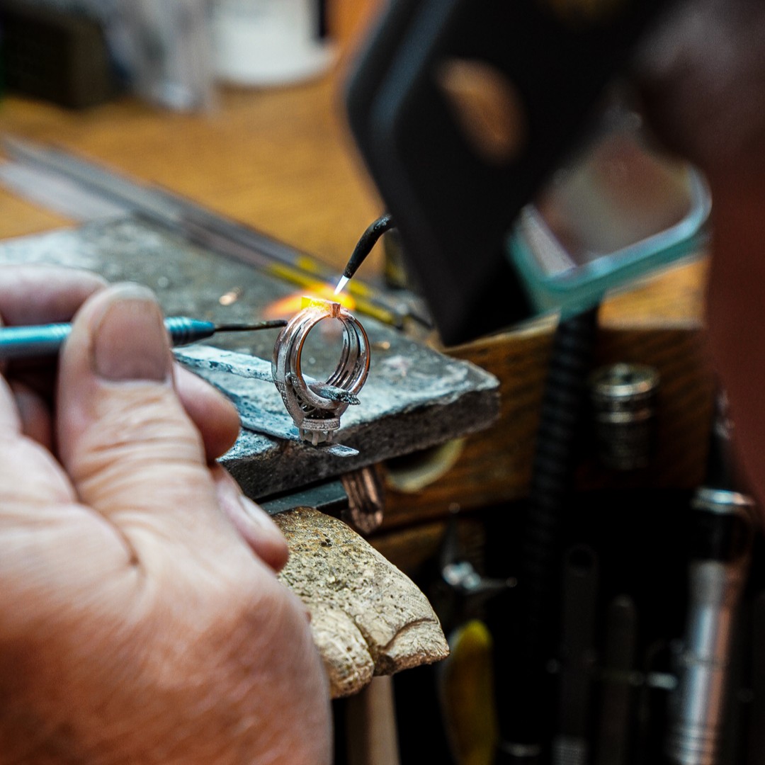 Jeweler using a small torch to repair a ring at a workbench with specialized jewelry tools.